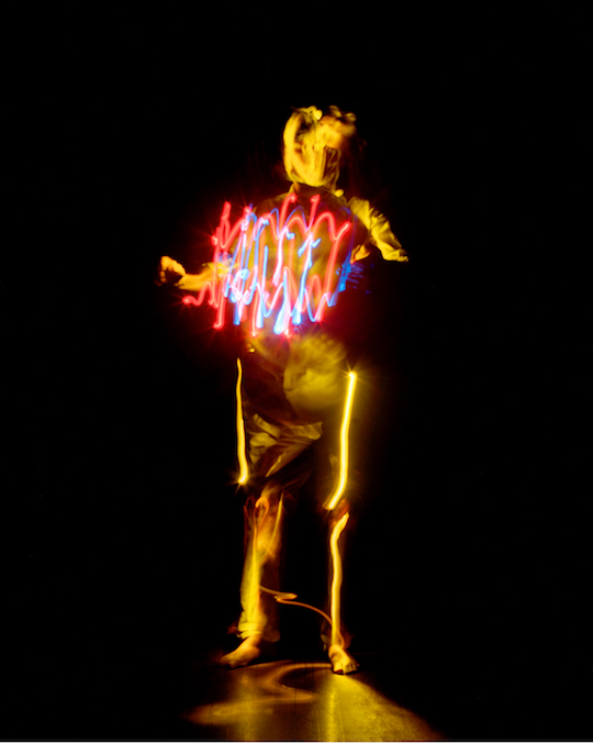 A photograph by Pete Eckert: A bright yellow figure in the middle of black, vaguely resembling a human body. The left arm seems to be cut in the middle, the right hand is stretched out as a fist. Head and face are barely recognizable, made out of yellow lightstreaks. In front of the upper body, in the hight of the heart, there are bright red and blue vibrant lines, resembling those of an oscillograph. Along the legs very bright yellow vertical lines as if this light would origin from the bones in the legs.