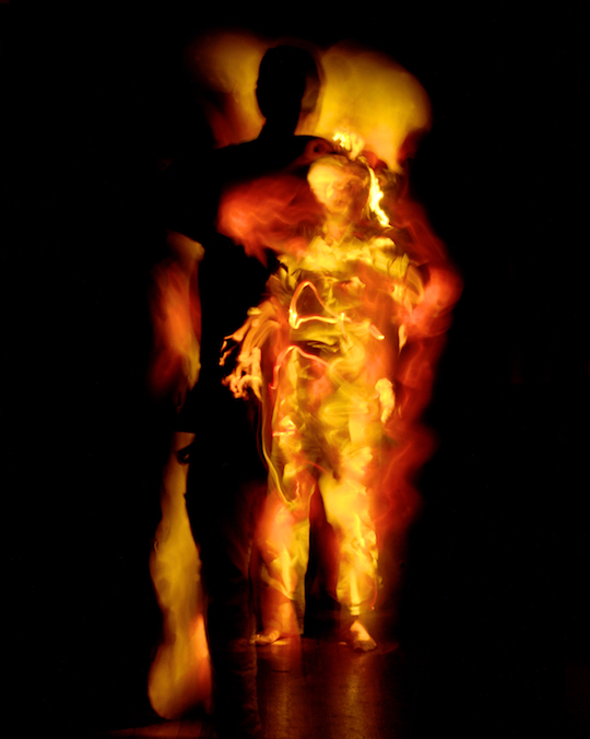 A photograph by Pete Eckert: Fiery orange-yellow streaks glowing out of the black. In the foreground stands a figure of vaguely human shape whose definitions are hard to make out, a strange head over a robe made out of lightstreaks. Only the left hand and the bare feet resemble cleary those of human bodies. Behind the figure a huge black shadow, dark and sharply carved out of the fiery lights. It is much taller than the figure who seems to cast it, towering over it.