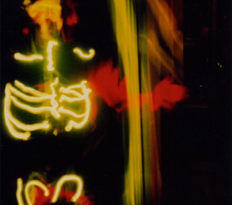 A photograph by Pete Eckert: In the left corner of the image there is a skeleton, greenish and yellow shining bones. On top of it something like a skincoloured head, the face hardly recognizable. On the right side of the skeleton strong vertical lines, painted with yellow-greenish and orange light, a separation in the image, appearing like a wall or a barrier. The skeleton reaches out with an arm out of red light like feathers, reaching through the vertical lines towards the other side of the image. On this side, the right side, there is black. And in the black some small lights, like shimmering out of a distance. The hand stays streched out, within this blackness.
